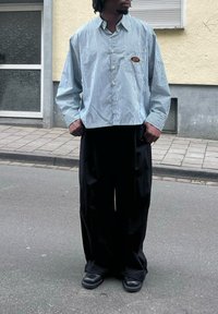 Camicia a righe blu e bianche, con bottoni, dalla vestibilità rilassata, dotata di una tasca sul petto con logo. I pantaloni neri larghi e le calzature nere completano l'outfit.
