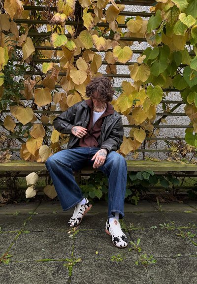 Chaqueta marrón con cremallera sobre una camisa blanca, jeans de pierna ancha azules y zapatos de estampado de vaca en blanco y negro. Sentado en un banco de madera, rodeado de hojas de otoño.