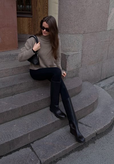 Mujer con gafas de sol, suéter beige de cuello alto, leggings negros y botas negras hasta la rodilla, sentada en escalones de piedra sosteniendo un bolso negro.