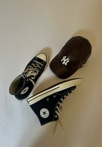 Tenisi negri cu glezna înaltă Converse, cu șireturi și tălpi albe, alături de o șapcă din piele maro închis a echipei New York Yankees, așezate pe o suprafață deschisă.