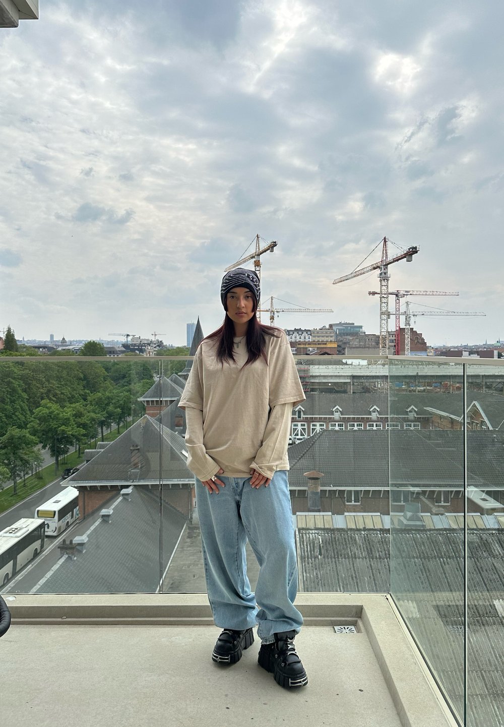 Beige oversized long-sleeve shirt, lichtblauwe baggy jeans en zwarte chunky sneakers. Buitenlocatie met kranen en een bewolkte lucht op de achtergrond.
