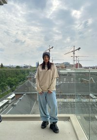 Beige oversized long-sleeve shirt, lichtblauwe baggy jeans en zwarte chunky sneakers. Buitenlocatie met kranen en een bewolkte lucht op de achtergrond.