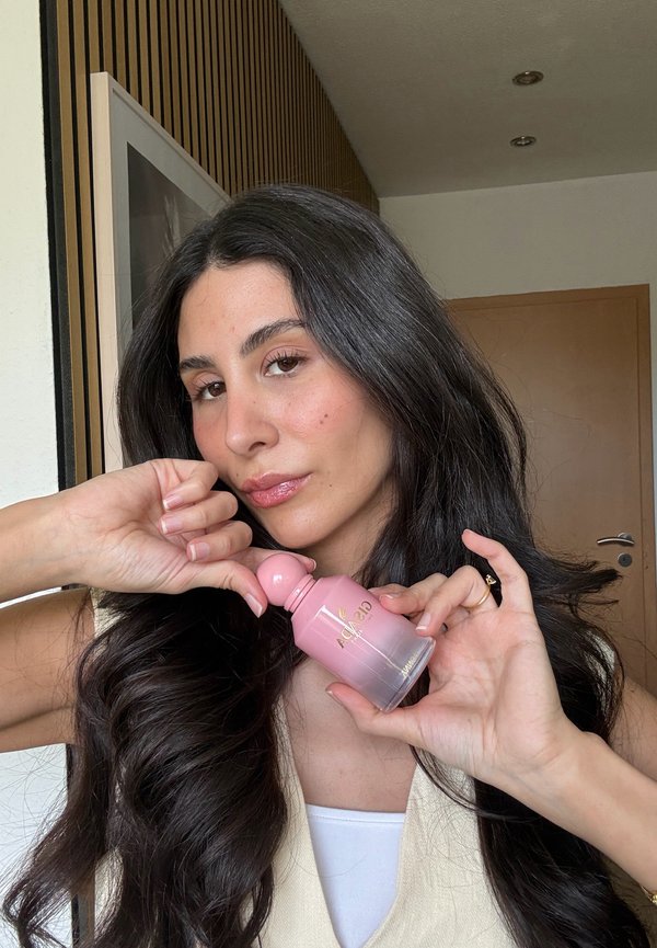 Femme aux longs cheveux foncés tenant un flacon de parfum rose étiqueté "Adagio", posant à l'intérieur près d'une porte en bois et d'une œuvre murale.