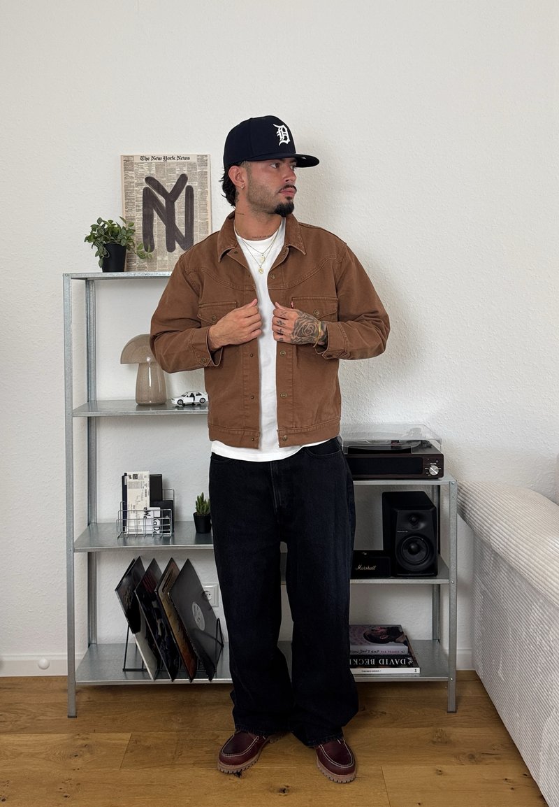 Veste en coton marron sur une chemise blanche, associée à un jean foncé et des chaussures marron. L'arrière-plan présente des étagères avec des décorations et des vinyles.