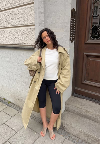 Mujer con cabello oscuro y rizado, vestida con gabardina beige, camiseta blanca, leggings negros recortados y sandalias beige, de pie junto a las escaleras de la entrada del edificio.