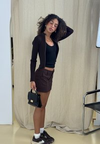 Brun zip-up cardigan över en svart tanktop, parat med bruna rutiga shorts och en svart handväska. Sneakers med vita strumpor. Neutral bakgrund.