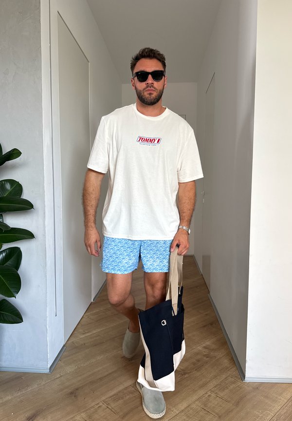 Homme portant des lunettes de soleil, un t-shirt blanc Tommy, un short bleu à motifs, et des chaussures grises sans lacets, portant un sac tote noir et beige à l’intérieur.