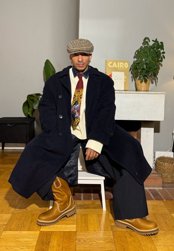 Bent ülő férfi, hosszú sötét kabátot, mintás nyakkendőt, fehér pulóvert, bő nadrágot, barna csizmát és kockás lapos sapkát visel.