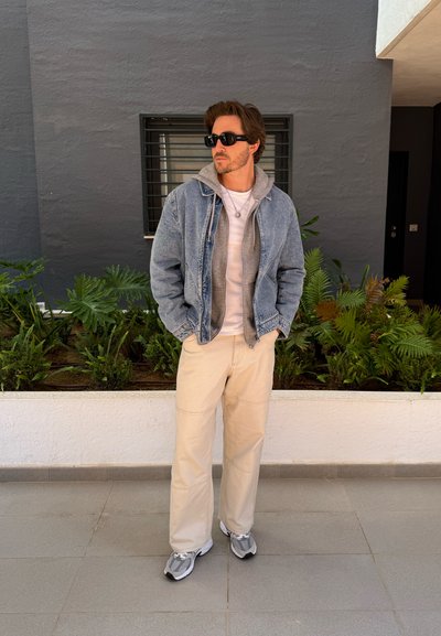Chaqueta de mezclilla de lavado claro sobre una sudadera gris, camisa blanca y pantalones beige de corte relajado. Zapatillas deportivas grises y gafas de sol negras.