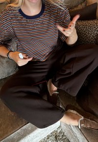 Chemise à manches courtes rayée en marron, crème et marine ; pantalon noir décontracté ; chaussons marron avec doublure en fausse fourrure ; tenant un stylo blanc.
