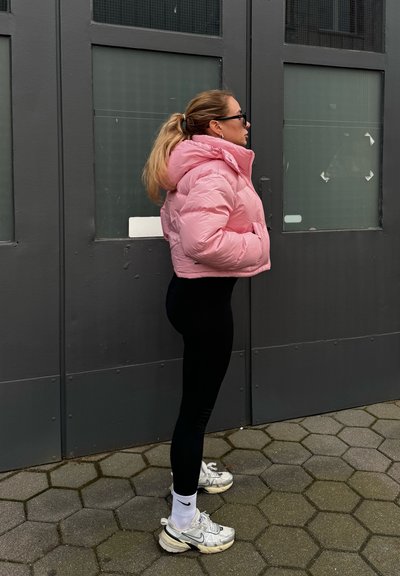 Chaqueta acolchada rosa con capucha, leggins negros y zapatillas deportivas plateadas. El cabello está peinado en una coleta, con gafas de sol y pendientes de aro.