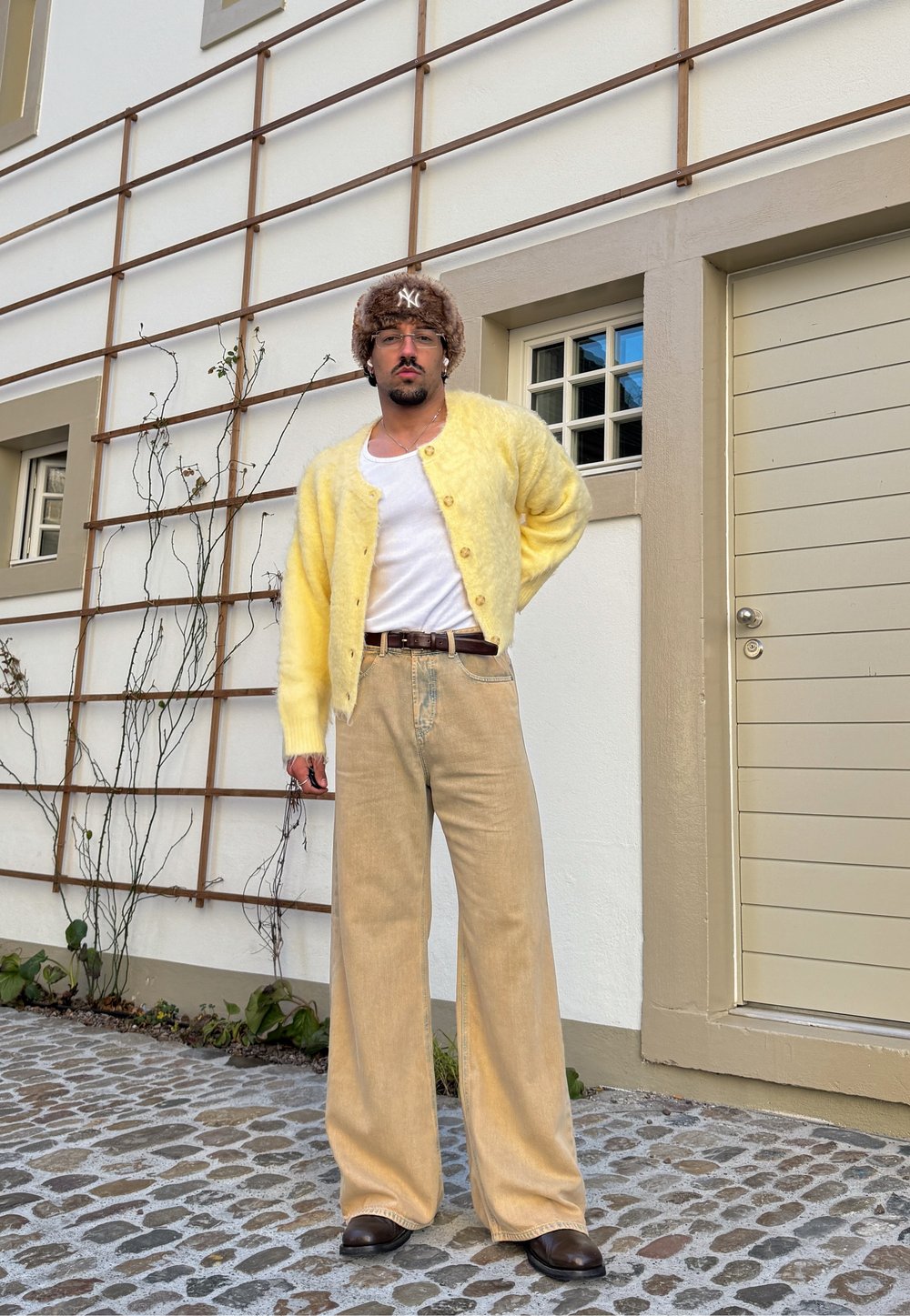 Άνδρας που φοράει χνουδωτή κίτρινη ζακέτα, λευκό αμάνικο μπλουζάκι, μπεζ παντελόνα, καφέ παπούτσια, χνουδωτό καπέλο και γυαλιά, στέκεται σε λιθ όστρωτο δρόμο.
