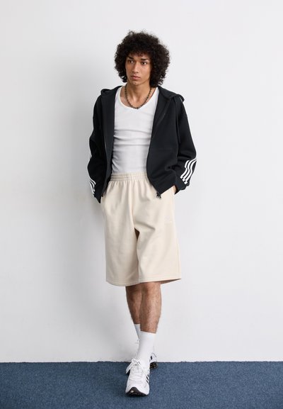 Sudadera negra con rayas blancas en las mangas, top blanco, pantalones cortos beige, calcetines blancos y zapatos deportivos. Fondo simple.