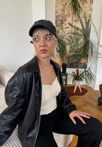Jachetă din piele neagră, top alb texturat, pantaloni negri și șapcă neagră. În fundal, plante și un tapet cu un model.