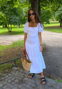 Fehér, rövid ujjú ruha négyzet alakú dekoltázzsal és réteges alsó szegéllyel. Fekete szandálok és barna fogantyúval ellátott szőtt táska. Kültéri környezet.