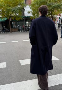 Dlhý tmavý vlnený kabát, nosený cez hnedé manžestrové nohavice. Osoba stojí na priechode pre chodcov s stromami a obchodmi v pozadí.