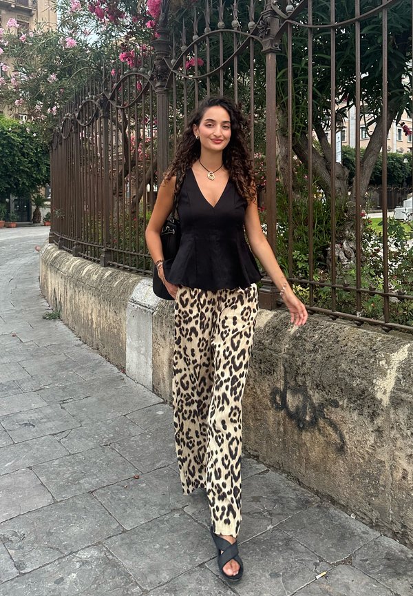 Čierny peplum top, béžové a čierne nohavice so zvieracou potlačou a širokými nohavicami, a čierne sandále na remienky. Exteriérové prostredie s kvetinovým pozadím.