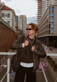 Svart skinnjakke over en mørk genser, kombinert med synlige hvite skjortemansjetter. Tilbehør inkluderer lagdelte sølvsmykker og svarte solbriller.