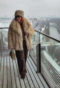 Femme en manteau et chapeau en fourrure beige, debout sur un balcon enneigé, surplombant une rivière et le paysage urbain par une journée d'hiver.
