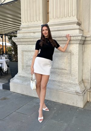 Top negro de manga corta, falda corta blanca, bolso blanco y sandalias blancas de tacón abierto. La modelo posa contra una pared de piedra con columnas acanaladas.