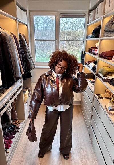 Persona con cabello rizado y gafas, vestida con chaqueta y pantalones de cuero marrón, de pie en un vestidor con ropa y estantes a ambos lados.