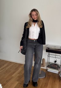 Zwarte blazer over een witte cropped blouse met knopen, gecombineerd met hoge taille grijze jeans en zwarte chunky schoenen. Houdt een zwarte tas vast. Vloer is van hout.