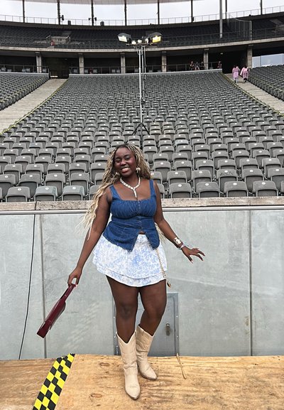 Crop top de mezclilla con peplum y falda corta de rayas azules y blancas; lleva botas de cowboy beige, sostiene un bolso burdeos, en un estadio vacío.