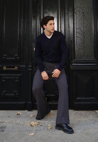 Granatowy sweter w serek, szare rozszerzane spodnie i czarne buty. Siedzi oparty o czarne drzwi z ozdobnym szklanym panelem.