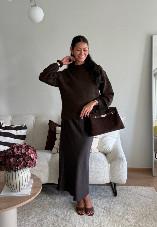 Une femme souriante porte un pull brun foncé, une jupe longue et un sac à main assorti dans un salon lumineux.