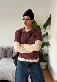 Άνδρας με γυαλιά ηλίου και μαύρο σκουφί, φορώντας πολλαπλά στρώματα μπλουζών και μπλε τζιν, στέκεται με σταυρωμένα τα χέρια δίπλα σε μια γλάστρα μέσα σε εσωτερικό χώρο.