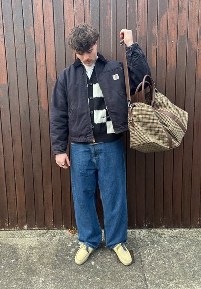 Chaqueta negra corta sobre un suéter de rayas blancas y negras, jeans azules holgados y zapatos beige. Se sostiene un bolso marrón a cuadros.
