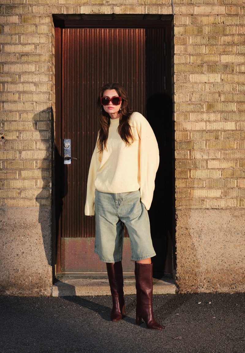 Femme portant un pull crème surdimensionné, un short en jean mi-cuisse, de grandes bottes bordeaux et de grandes lunettes de soleil, se tenant contre un mur de briques et une porte sombre.