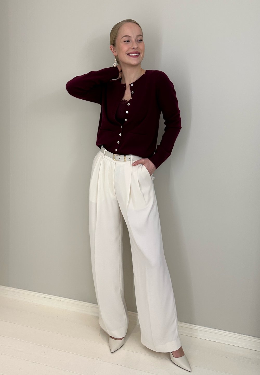 Cardigan bordeaux avec boutons, pantalon blanc plissé et chaussures pointues beige clair. Accessoires comprenant de grandes boucles d'oreilles pendantes. Textures lisses.