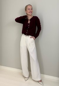 Cardigan bordeaux avec boutons, pantalon blanc plissé et chaussures pointues beige clair. Accessoires comprenant de grandes boucles d'oreilles pendantes. Textures lisses.