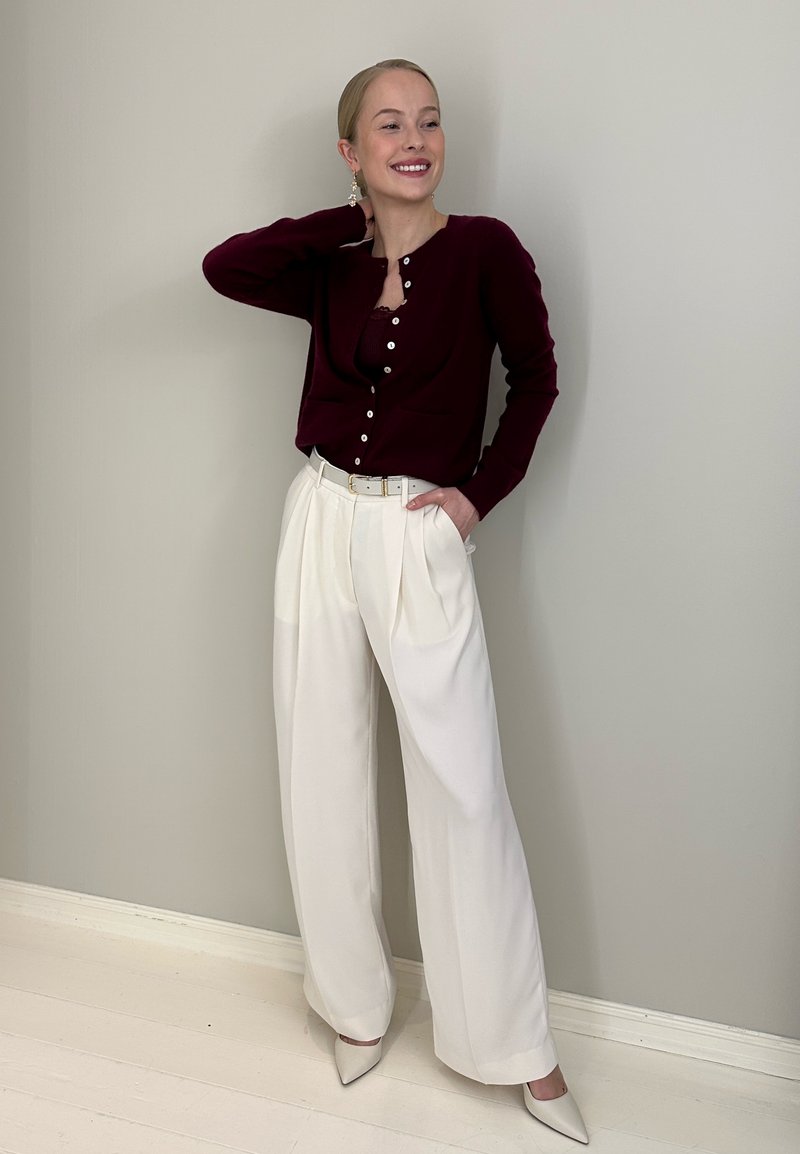 Cardigan bordeaux avec boutons, pantalon blanc plissé et chaussures pointues beige clair. Accessoires comprenant de grandes boucles d'oreilles pendantes. Textures lisses.
