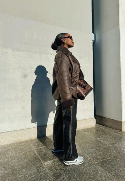 Chaqueta de cuero marrón con forro de forro polar, pantalones negros y zapatillas plateadas. El accesorio incluye un bolso de cuadros marrón. Sombra visible en la pared.