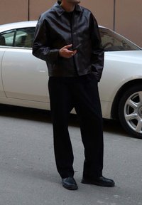 Chaqueta corta de cuero marrón con cierre de botón, combinada con pantalones de pierna ancha negros y zapatos negros, de pie junto a un coche blanco.