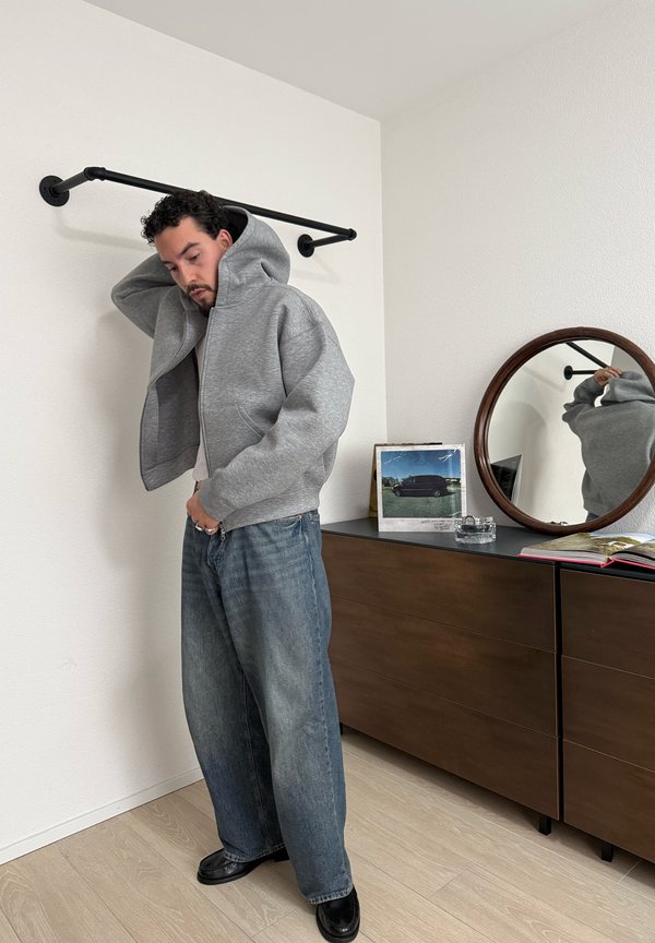 Mand iført grå hættetrøje og løse jeans står ved et trækammerskab med et rundt spejl og indrammet foto indendørs.