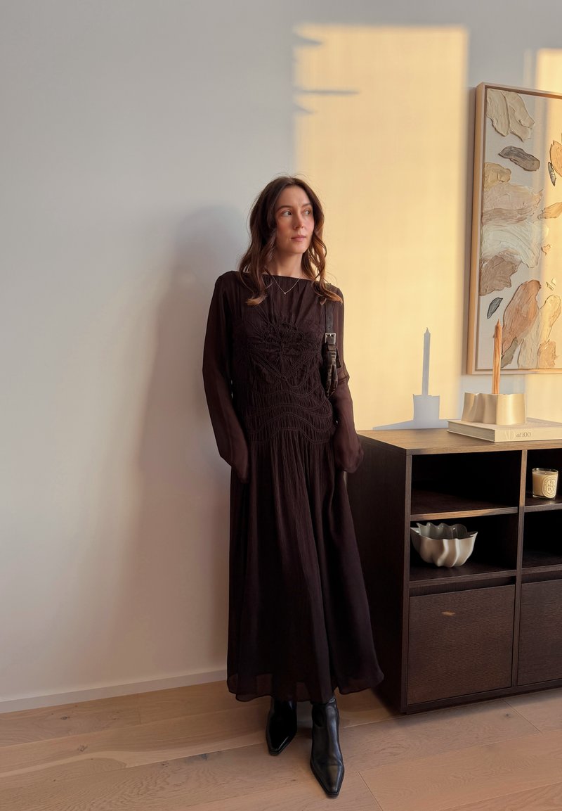 Femme en robe longue noire et bottes appuyée contre un mur près d'une armoire en bois avec décoration et peinture abstraite dans une douce lumière du soir.