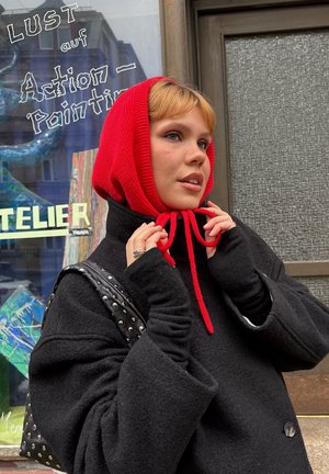 Joven con bufanda roja con capucha y abrigo negro ajustando la capucha cerca de una tienda con texto pintado y materiales de arte.