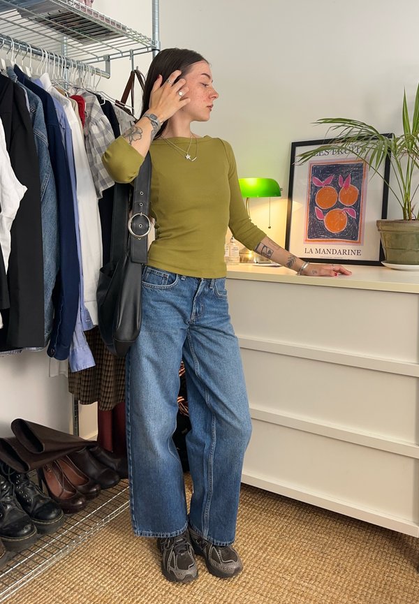 Vrouw in groen shirt en wijde spijkerbroek staat bij een witte commode en kledingrek, raakt haar haar aan en leunt op de commode met een ingelijst kunstwerk in de buurt.