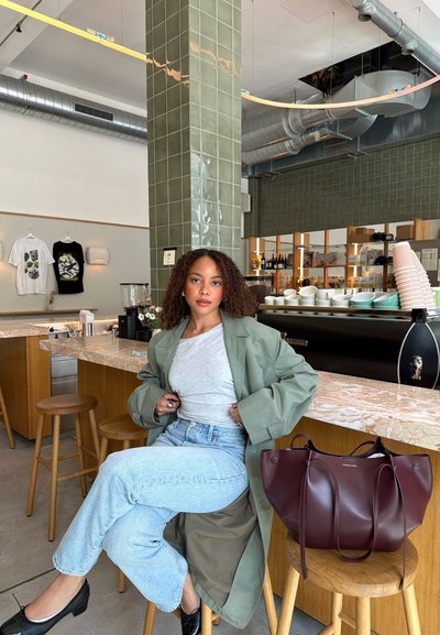 Mujer con cabello rizado, abrigo verde y jeans sentada en un taburete en el mostrador de una cafetería con tazas, bolsas y camisas exhibidas al fondo.