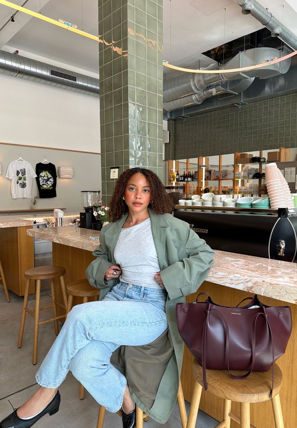 Donna con capelli ricci, cappotto verde e jeans seduta su uno sgabello al bancone di un bar con tazze, borse e camicie esposte sullo sfondo.