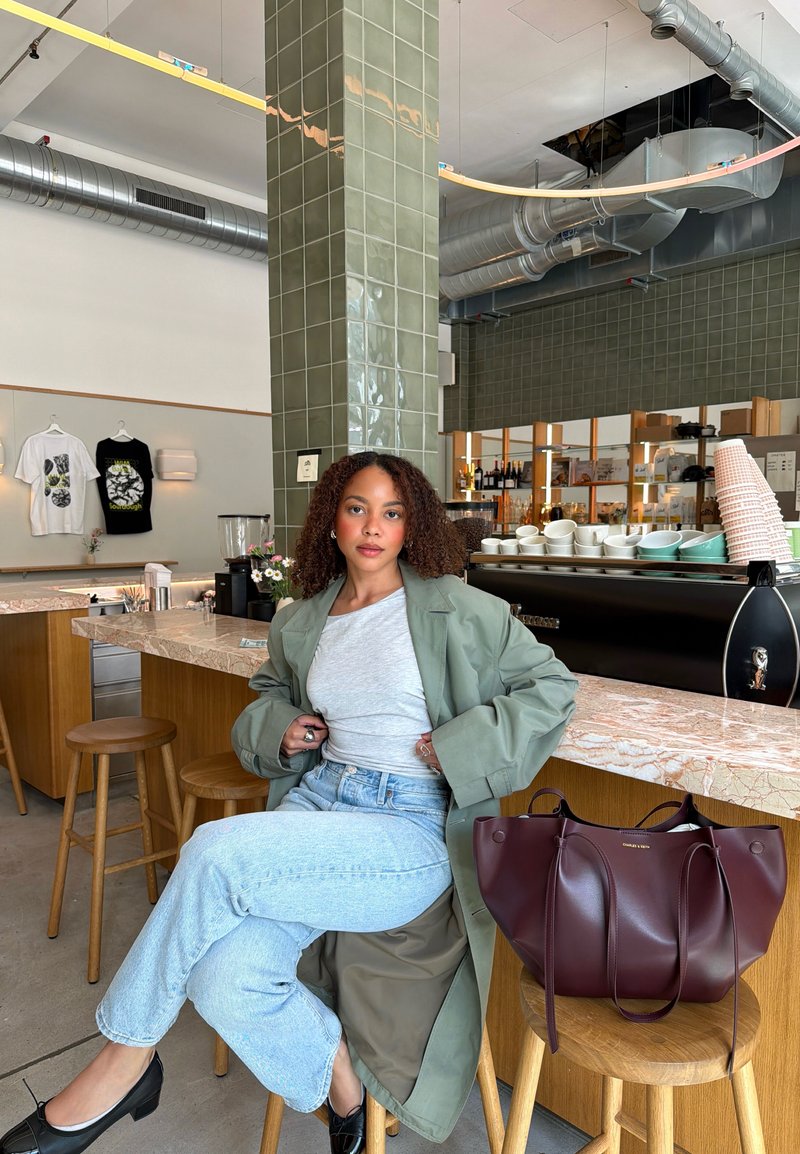 Femme aux cheveux bouclés, manteau vert et jeans, assise sur un tabouret au comptoir d'un café avec des tasses, sacs et chemises exposés en arrière-plan.