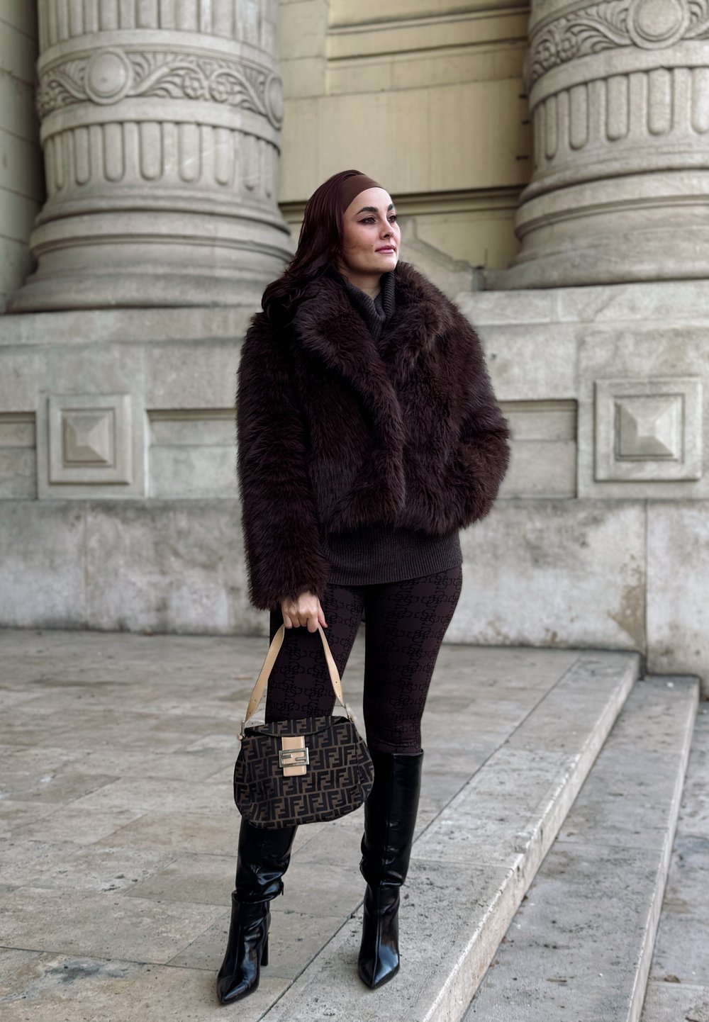 Καφέ ψεύτικη γούνα μπουφάν, εφαρμοστό σκουρόχρωμο πουλόβερ, μοτίβο κολάν, μαύρες ψηλές μπότες με στιλέτο, και υφασμάτινη τσάντα με χρυσό κούμπωμα.