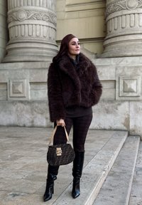 Καφέ ψεύτικη γούνα μπουφάν, εφαρμοστό σκουρόχρωμο πουλόβερ, μοτίβο κολάν, μαύρες ψηλές μπότες με στιλέτο, και υφασμάτινη τσάντα με χρυσό κούμπωμα.