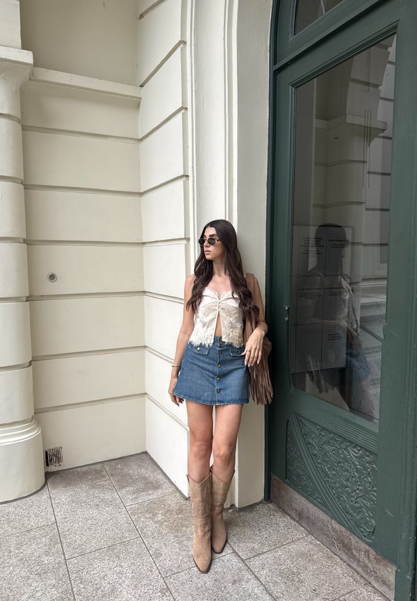 Femme aux longs cheveux foncés portant des lunettes de soleil, un top blanc sans manches, une jupe en jean et des bottes marron clair, debout près d'une porte verte sur un trottoir pavé de pierre.
