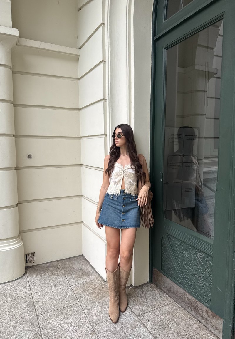 Femme aux longs cheveux foncés portant des lunettes de soleil, un top blanc sans manches, une jupe en jean et des bottes marron clair, debout près d'une porte verte sur un trottoir pavé de pierre.
