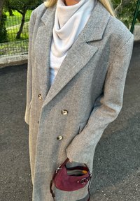 Manteau croisé en chevron gris avec de grands boutons dorés, porté sur un col roulé gris clair. Un petit sac à main bordeaux est tenu dans une main.