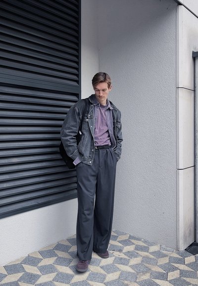 Chaqueta de mezclilla negra sobre una camisa lila clara, combinada con pantalones oscuros de tiro alto y zapatos burdeos, de pie sobre un suelo con patrón.